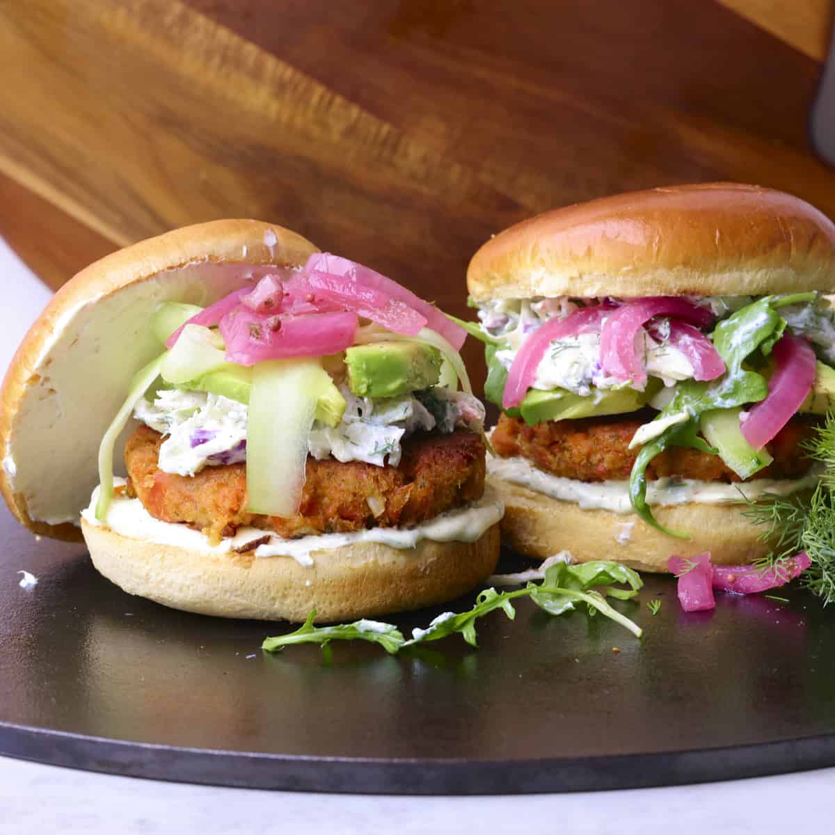 Two fully dressed Salmon Burgers with Canned Salmon, one with the top propped up against it and the other topped with the bun.