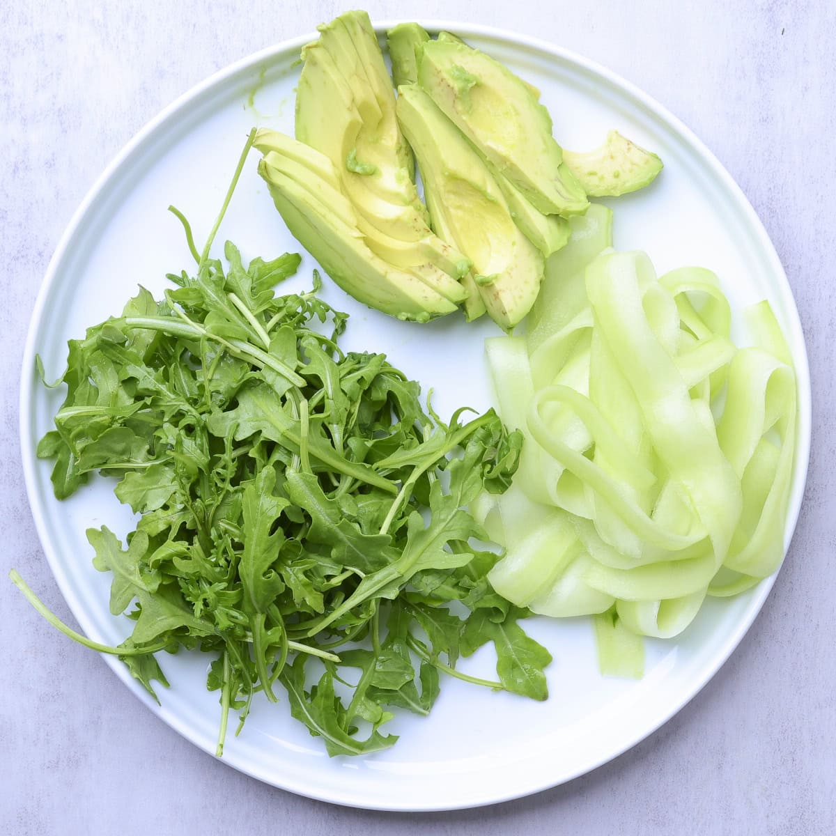 A plate of julienne cucumbers, sliced avocado and washed arugula.