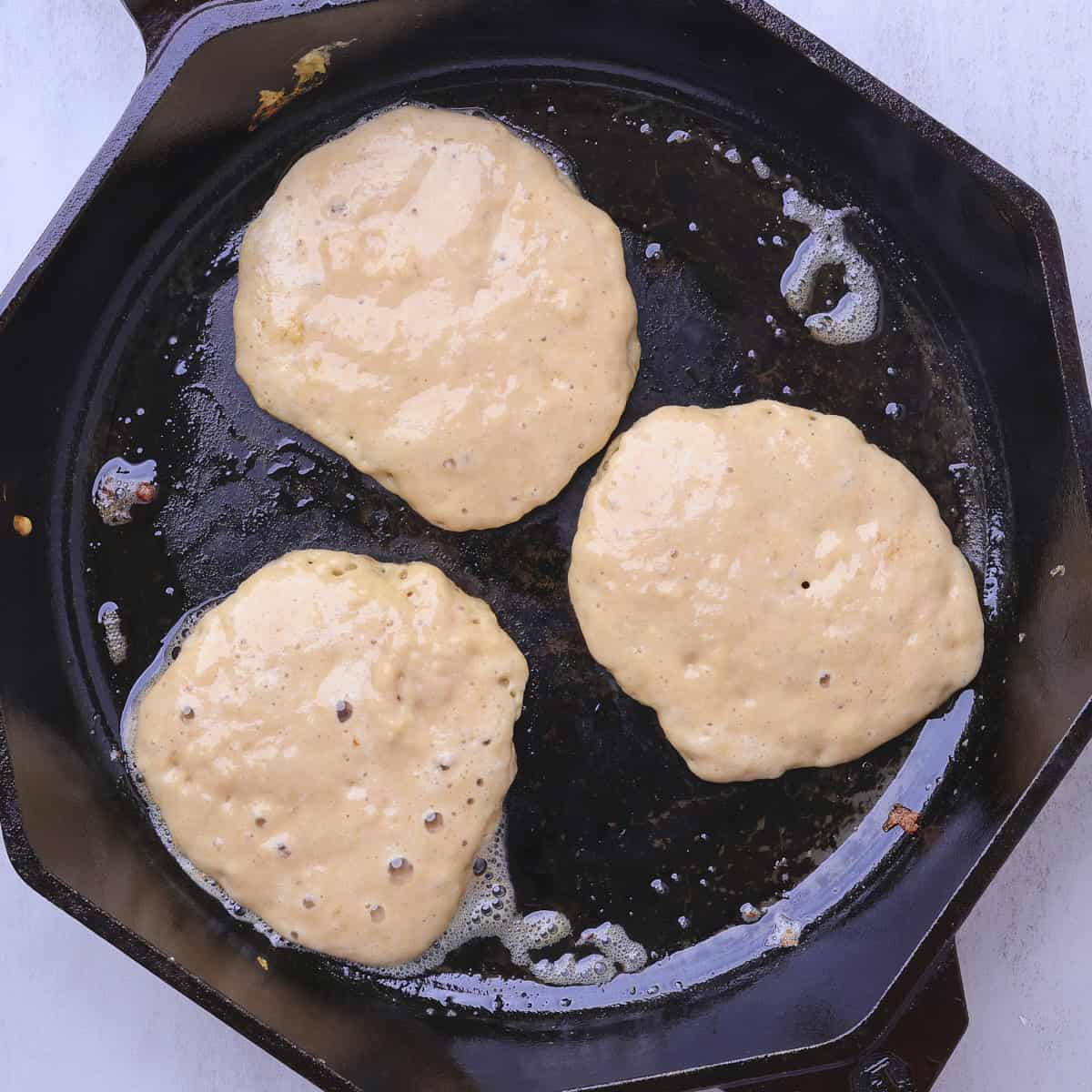 A hot skillet containing three sourdough banana pancakes, showing bubbles in the batter signifying they are ready to be flipped. 