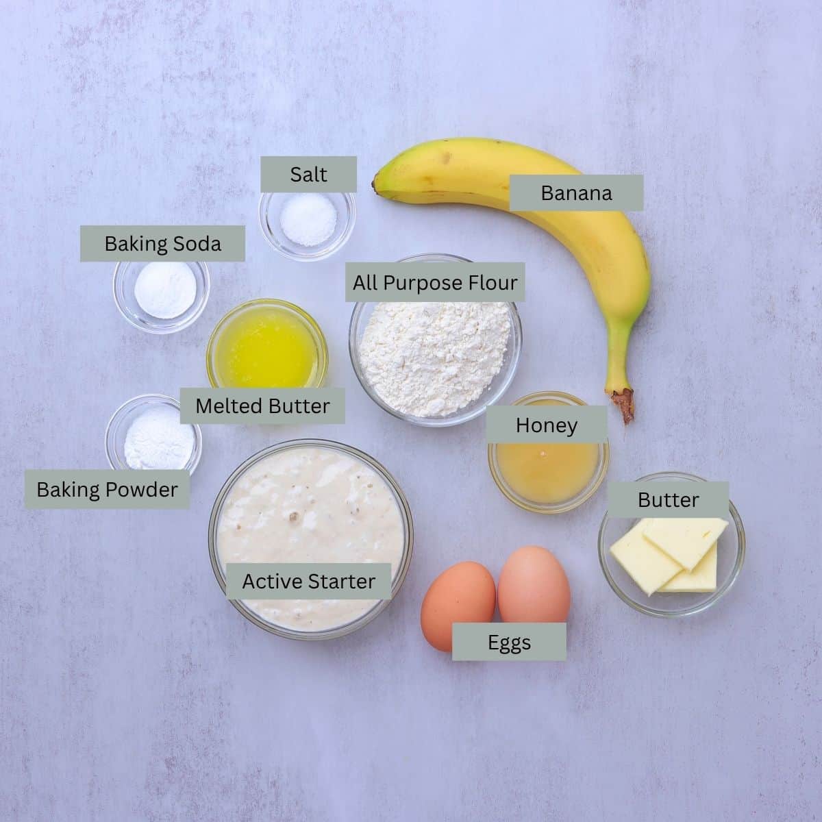  A visual display of the ingredients needed for Sourdough Banana Pancakes - banana, flour, salt, baking soda, baking powder, melted butter, honey, eggs, butter (for frying), and active sourdough starter - each in their own bowl.
