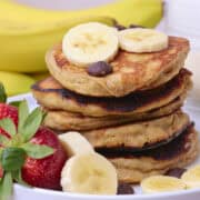 Sourdough Banana Pancakes Stacked on a plate and displayed with fruit and chocolate chips.