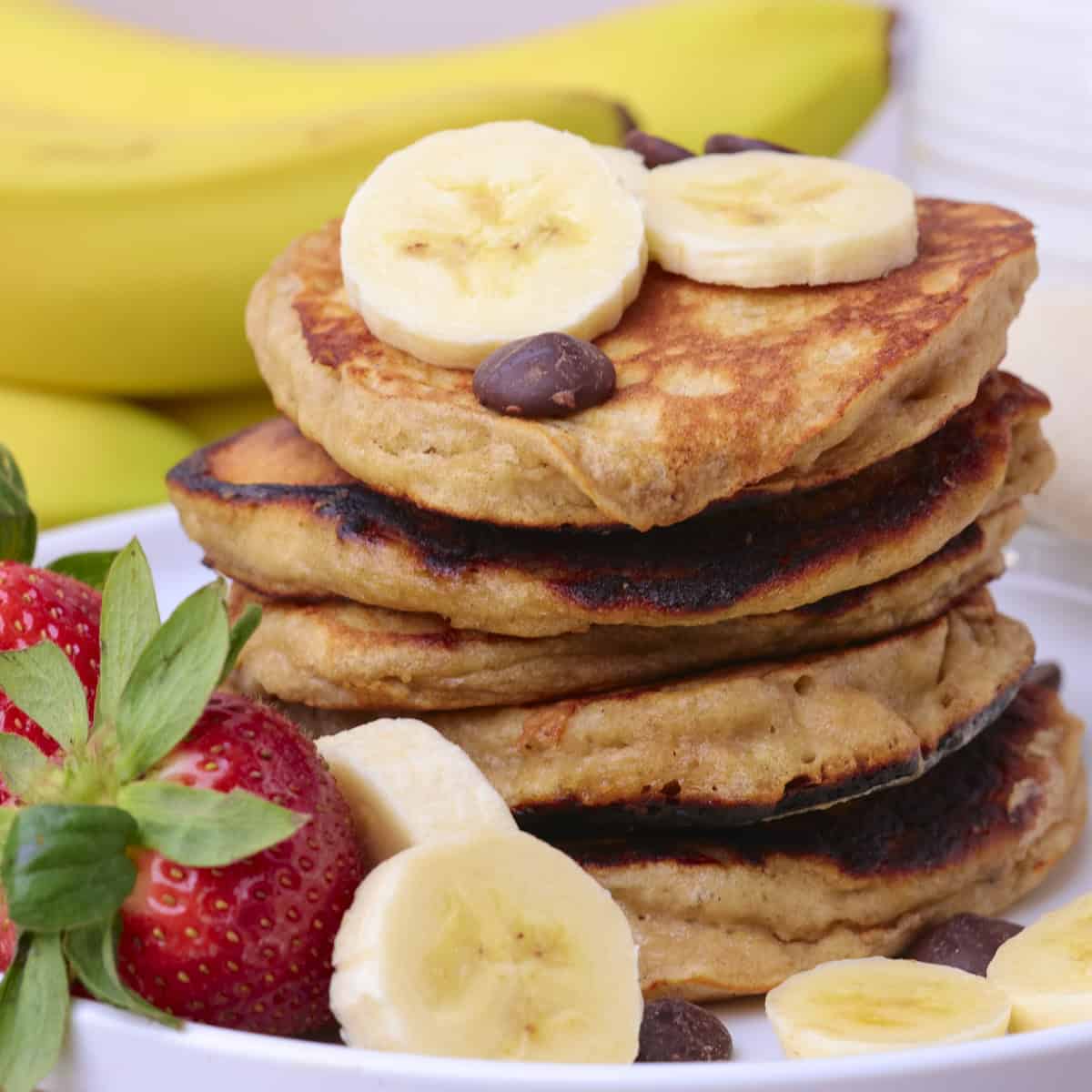 Sourdough Banana Pancakes Stacked on a plate and displayed with fruit and chocolate chips.