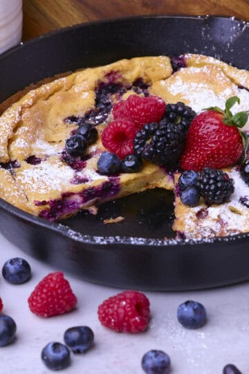A baked Sourdough German Pancake in a skillet with a slice removed, topped with icing sugar and berries.