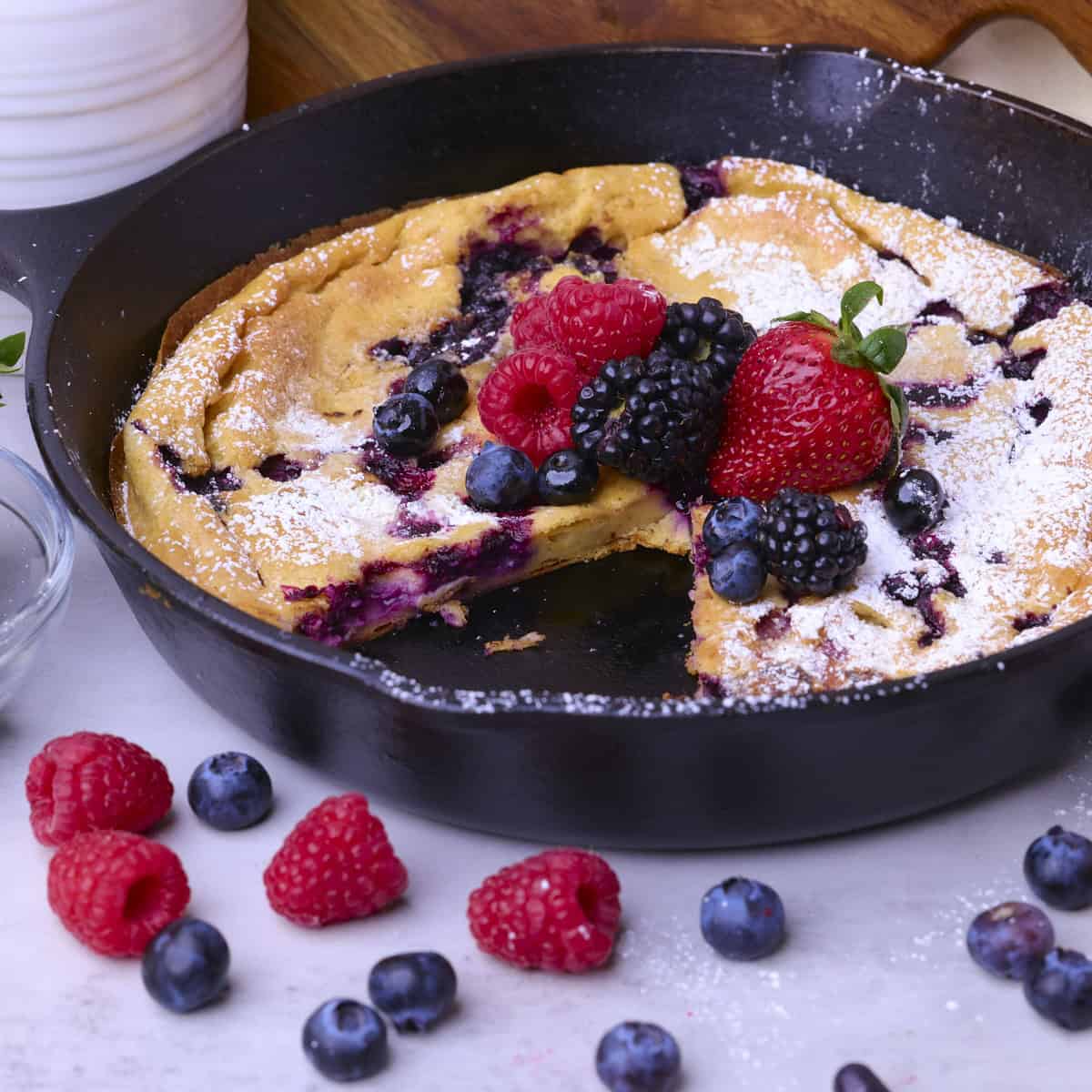 A baked Sourdough German Pancake in a skillet with a slice removed, topped with icing sugar and berries.