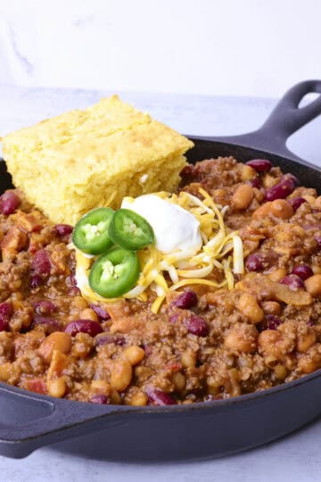 A raw cast iron skillet with Baked Beans with Hamburger and Bacon, topped with shredded cheese, sour cream and jalapeno slices with a piece of corn bread resting on top.