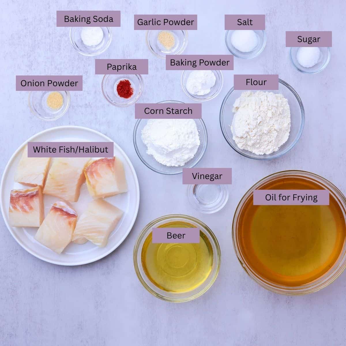 A visual display of the ingredients needed for Beer Battered Halibut - onion powder, paprika, garlic powder, salt, sugar, baking soda, baking powder, corn starch, flour, vinegar, beer, oil for frying and halibut - each displayed in their own bowl.