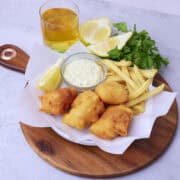 A wooden board wit ha plate of Beer Battered Halibut on parchment paper with fries, tartar sauce and a lemon wedge displayed with greens and a glass of beer.