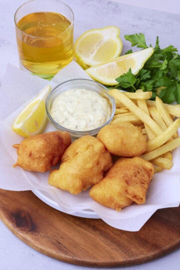 A wooden board wit ha plate of Beer Battered Halibut on parchment paper with fries, tartar sauce and a lemon wedge displayed with greens and a glass of beer.