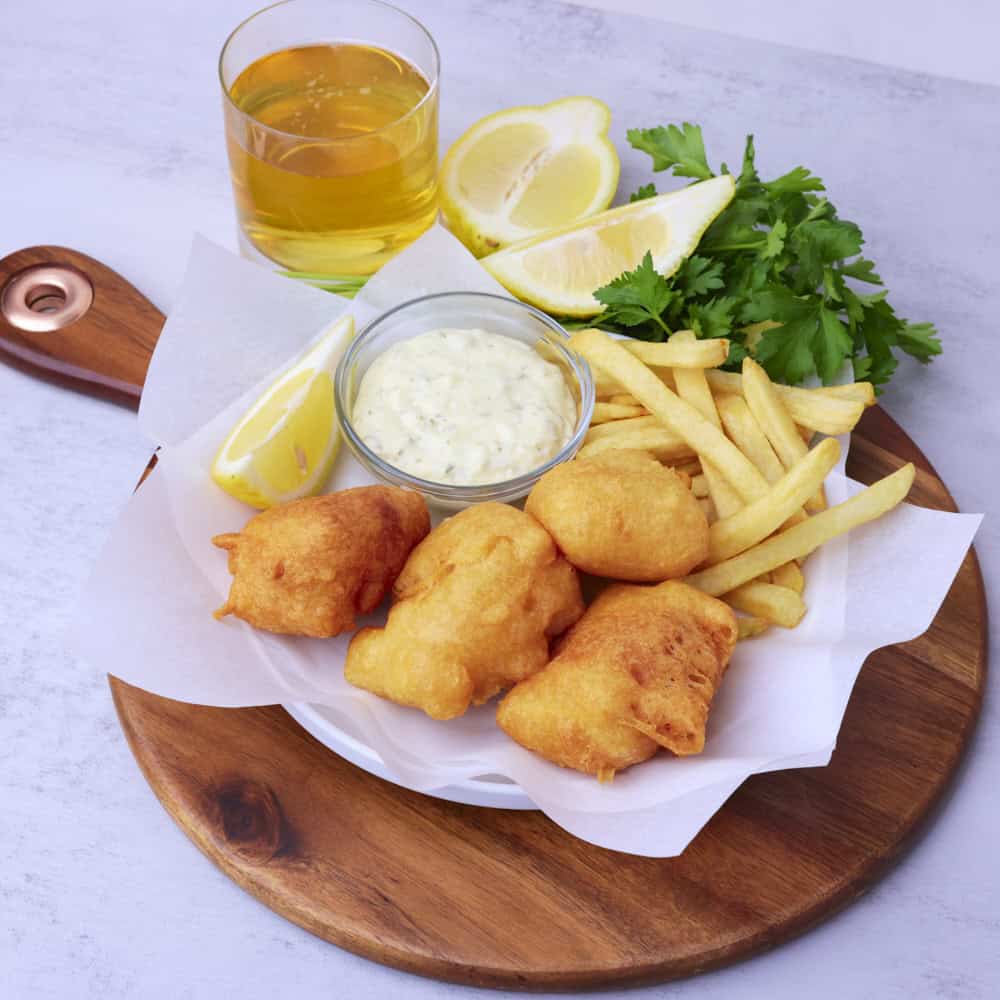 A wooden board wit ha plate of Beer Battered Halibut on parchment paper with fries, tartar sauce and a lemon wedge displayed with greens and a glass of beer.