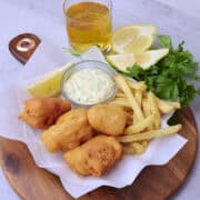 A wooden board wit ha plate of Beer Battered Halibut on parchment paper with fries, tartar sauce and a lemon wedge displayed with greens and a glass of beer.