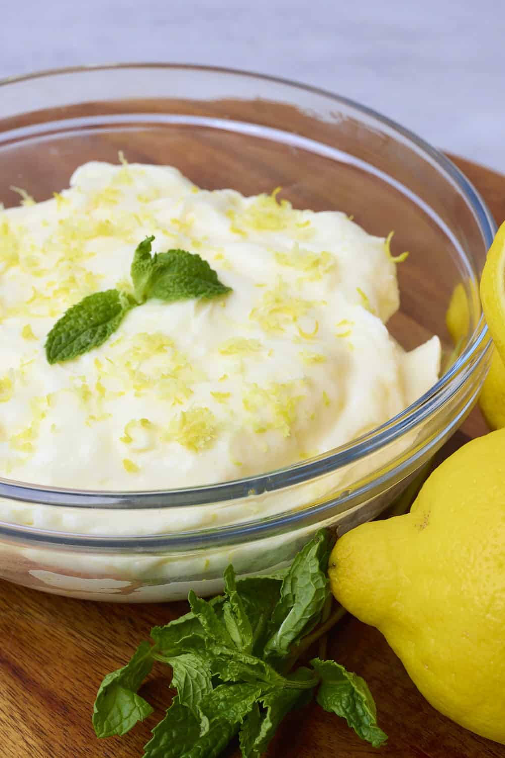 	A bowl of Lemon Cream Cheese Icing topped with zest and mint leaves placed displayed with lemons and mint beside.