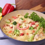 A pot of halibut pasta displayed against a wood board with greens and a red bell pepper.