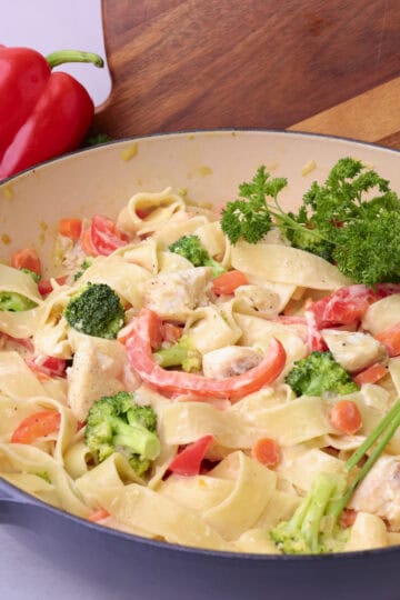 A pot of halibut pasta displayed against a wood board with greens and a red bell pepper.