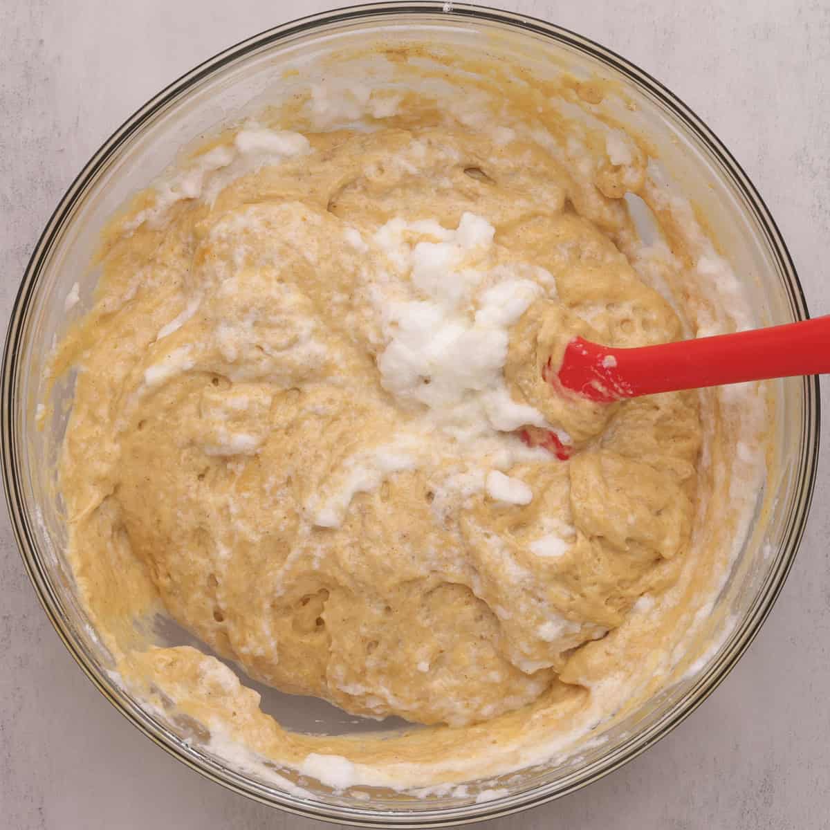 A glass bowl with the Sourdough Pumpkin Pancake batter mixed together showing a few streaks and lumps of egg whites showing with a red spatula.
