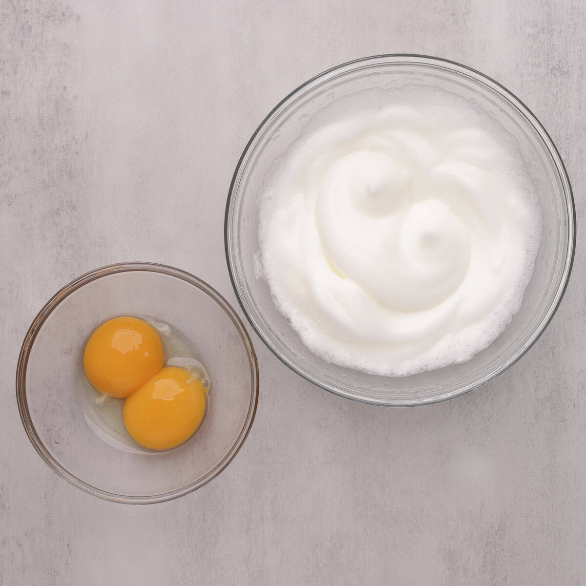 Two glass bowls, one containing two egg yolks and the other whipped egg whites.