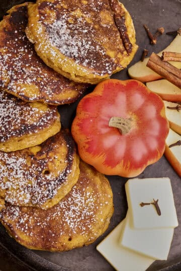 A skillet with Sourdough Pumpkin Pancakes in it displayed with a mini pumpkin, apple slices, cinnamon sticks and butter slices.