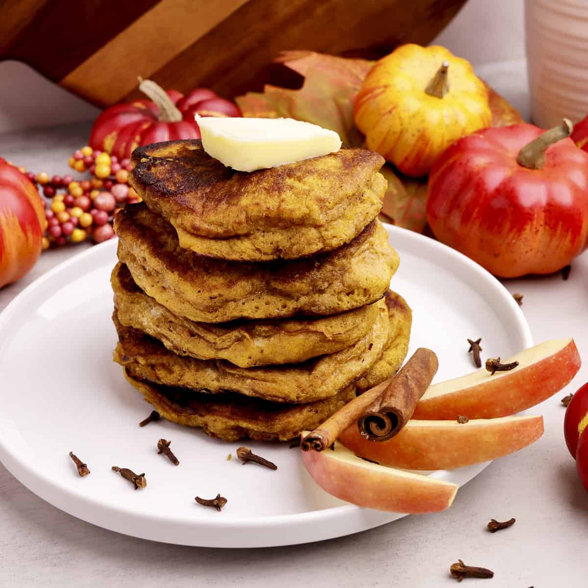 A stack of Sourdough Pumpkin Pancakes topped with butter on a white plate displayed with apple slices, cinnamon sticks, cloves and mini pumpkins.
