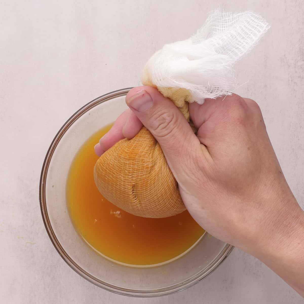 Hand holding cheesecloth filled with pumpkin puree and squeezing out the juices into a glass bowl beneath.