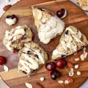An overhead view of a group of scones displayed on a wooden board with cherries and nuts sprinkled around.