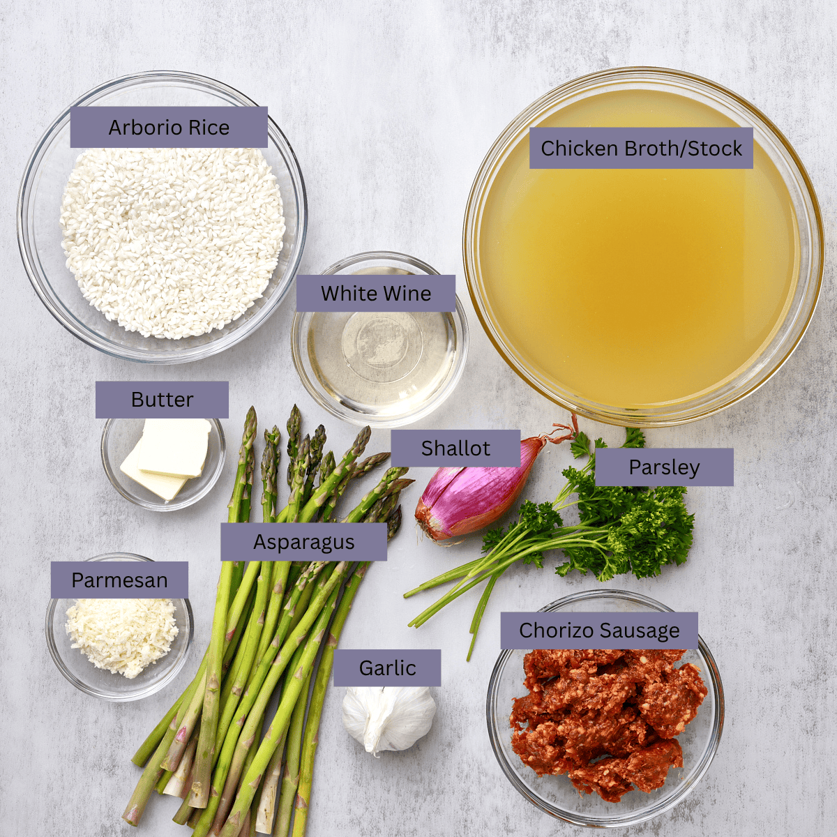 A visual display of the ingredients needed for the Chorizo Risotto - arborio rice, chicken broth, white wine, shallot, parsley, chorizo sausage, garlic, asparagus, butter and Parmesan - each displayed in their own bowl. 