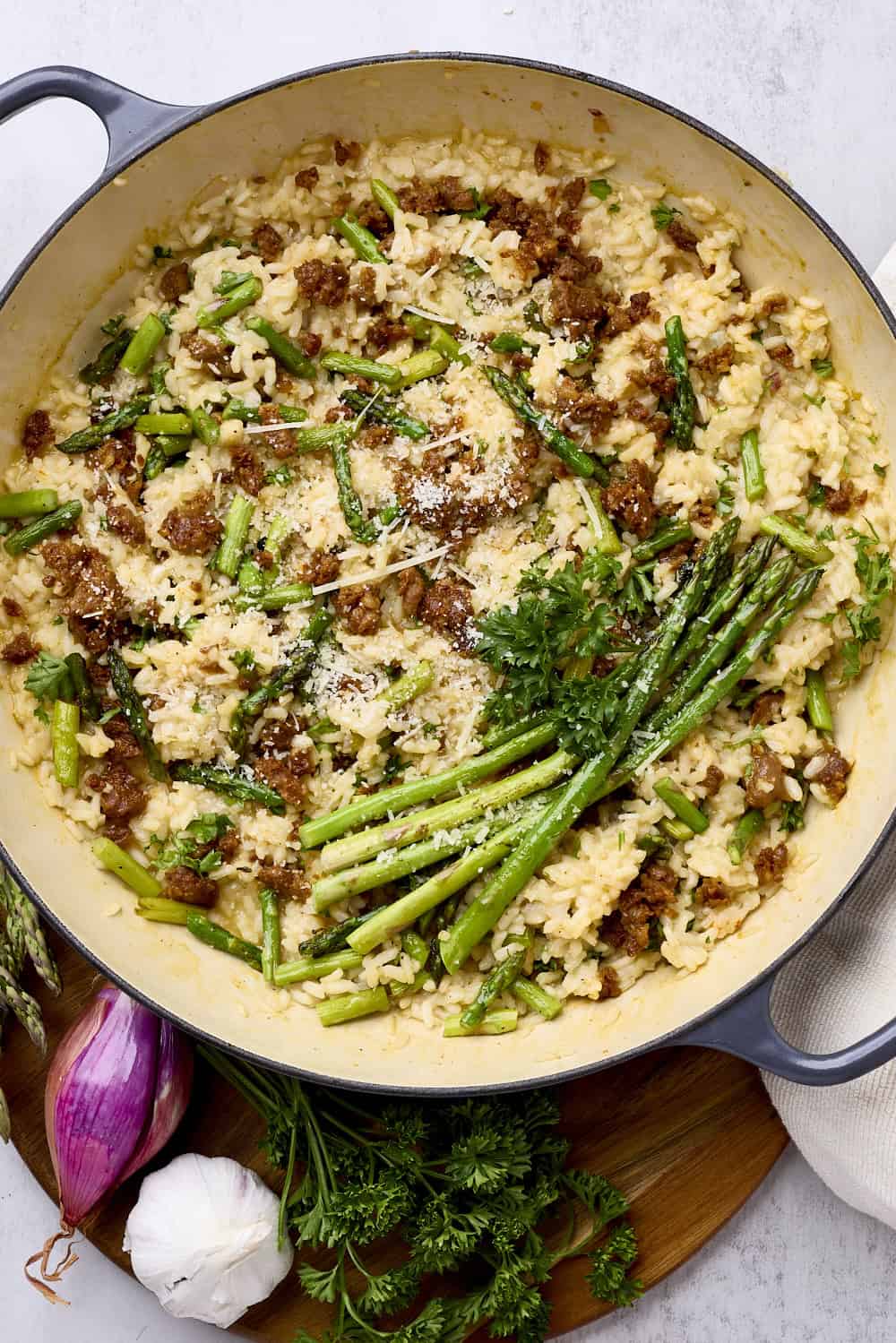 A pan filled with Chorizo Risotto topped with asparagus and displayed with a shallot, garlic head and greens.