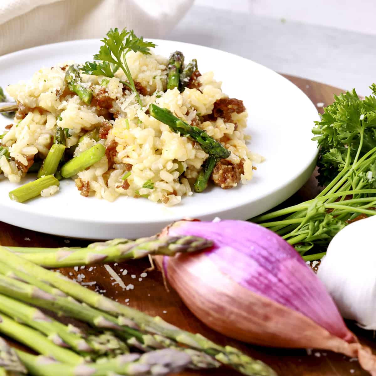 A white plate with a portion of Chorizo Risotto displayed with asparagus, a shallot, garlic and greens.