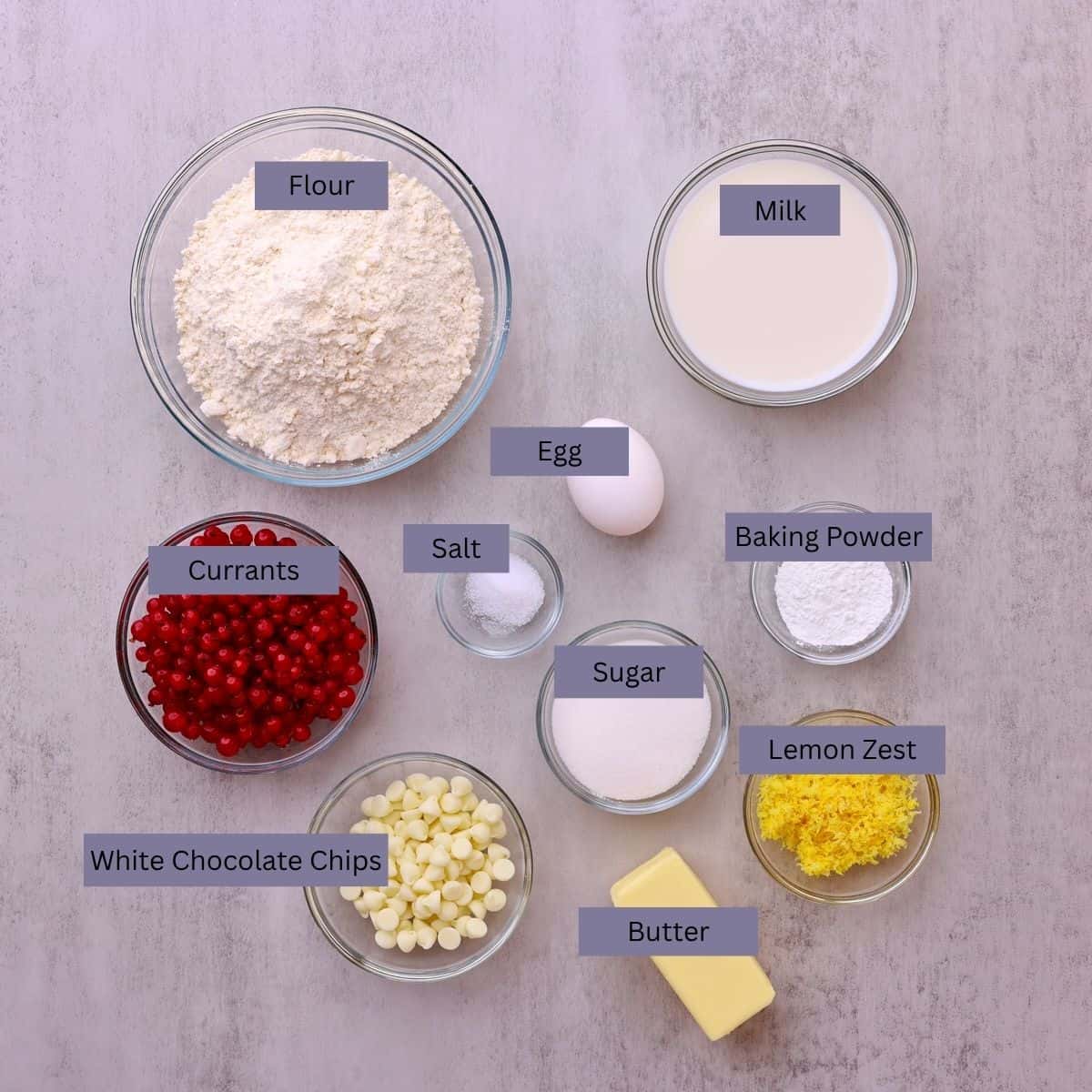 A visual display of the ingredients - flour, milk, currants, salt, egg, baking powder, sugar, white chocolate chips, butter and lemon zest - each displayed in their own bowl.