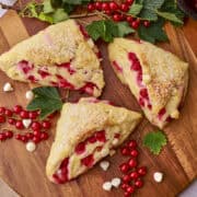 Three Currant Scones displayed on a wooden board with bunches of currants, greens and white chocolate chips.