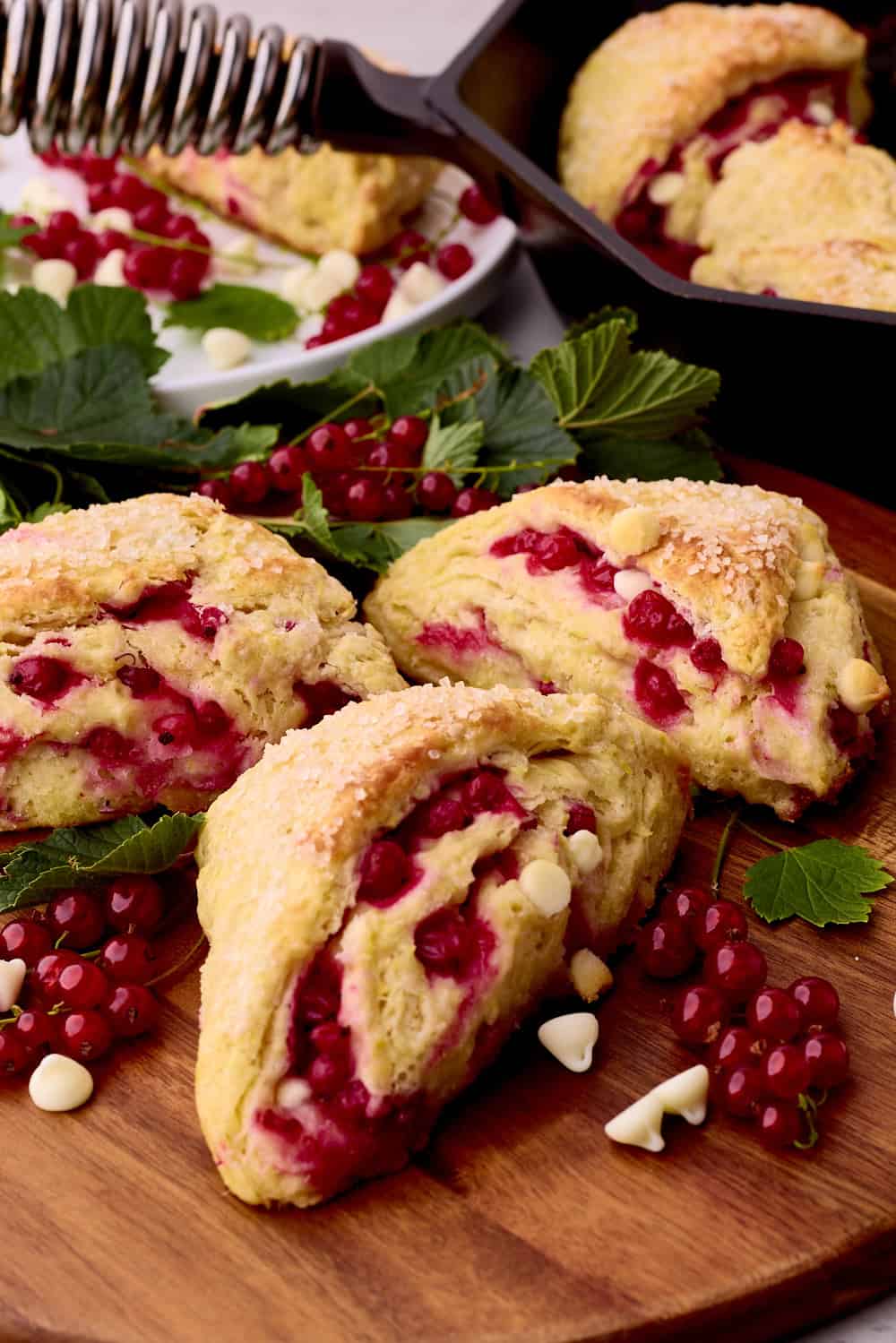 The Currant Scones displayed on a wooden board with brunches of currants, white chocolate chips and greens around them.