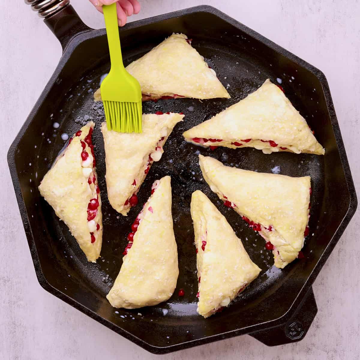 A skillet filled with the scones topped with milk and sugar with a brush touching a scone.