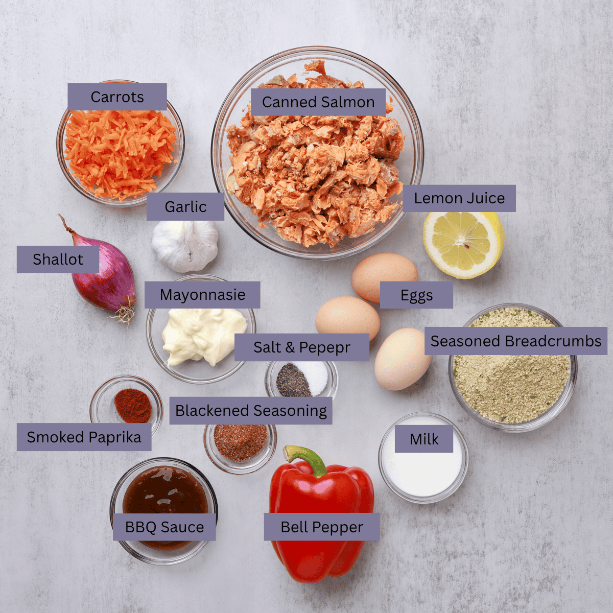 A visual display of the ingredients needed for the salmon loaf - canned salmon, carrot, shallot, garlic, lemon juice, eggs, mayonnaise, smoked paprika, barbecue sauce, blackened seasoning, salt, pepper, milk, bread crumbs and bell pepper - each displayed in their own bowl. 