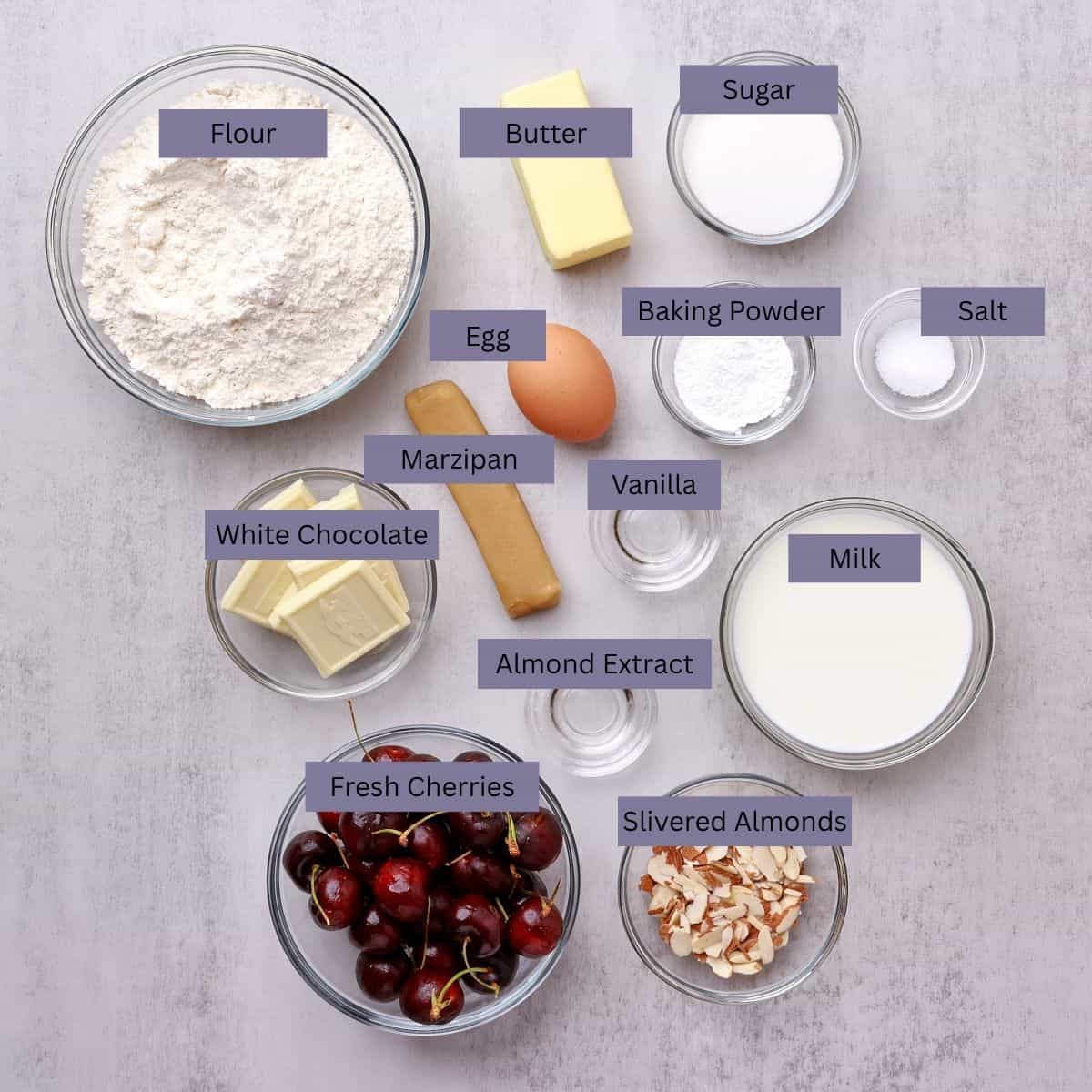 A visual display of the ingredients needed for the Cherry Scones - flour, butte,r sugar, marzipan, an egg, baking powder, salt, bakers white chocolate, vanilla, almond extract, milk, fresh cherries and slivered almonds - each displayed in their own bowl.