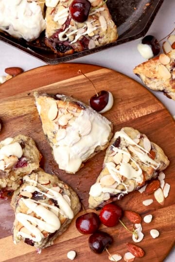 An overhead view of the Cherry Scones displayed on a wooden board with cherries and nuts, two with bites out of them and the skillet in the corner.