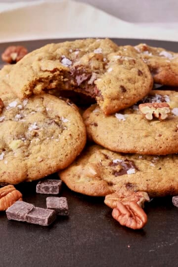 A pile of cookies on a cast iron platter sprinkled with chocolate chunks and pecans. The top cookie with a bite out of it.