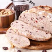 A loaf of bread with two slices in front, displayed on a wooden board with walnuts and craisins.