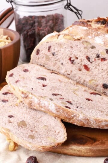 A loaf of bread with two slices in front, displayed on a wooden board with walnuts and craisins.