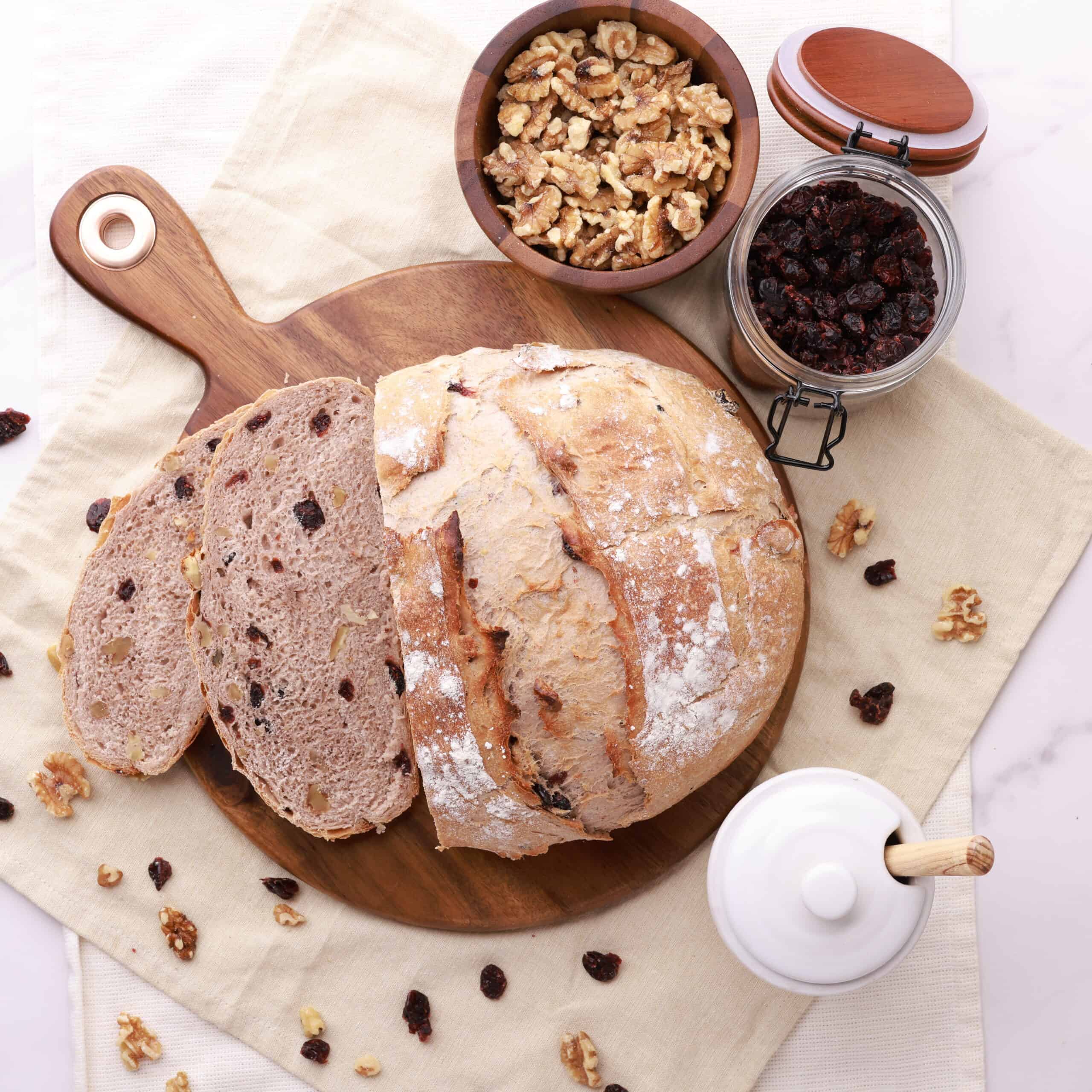 An overhead shot of the loaf with two slices, displayed on a wooden board with craisins and walnuts sprinkled around and in jars beside.
