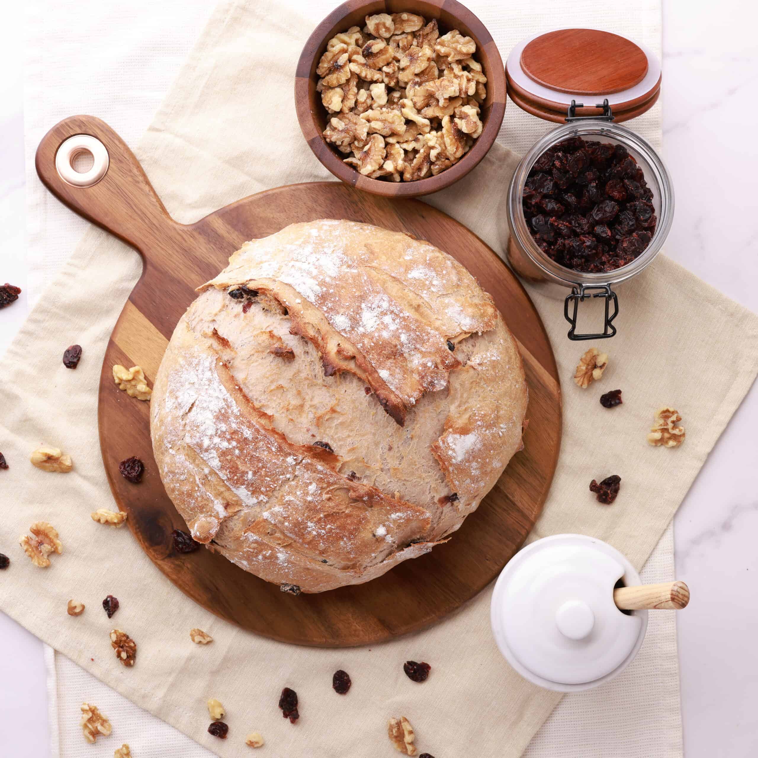 An overhead shot of the whole, un-sliced cranberry walnut loaf displayed on a cream cloth with walnuts and craisins sprinkled around and in bowls beside.
