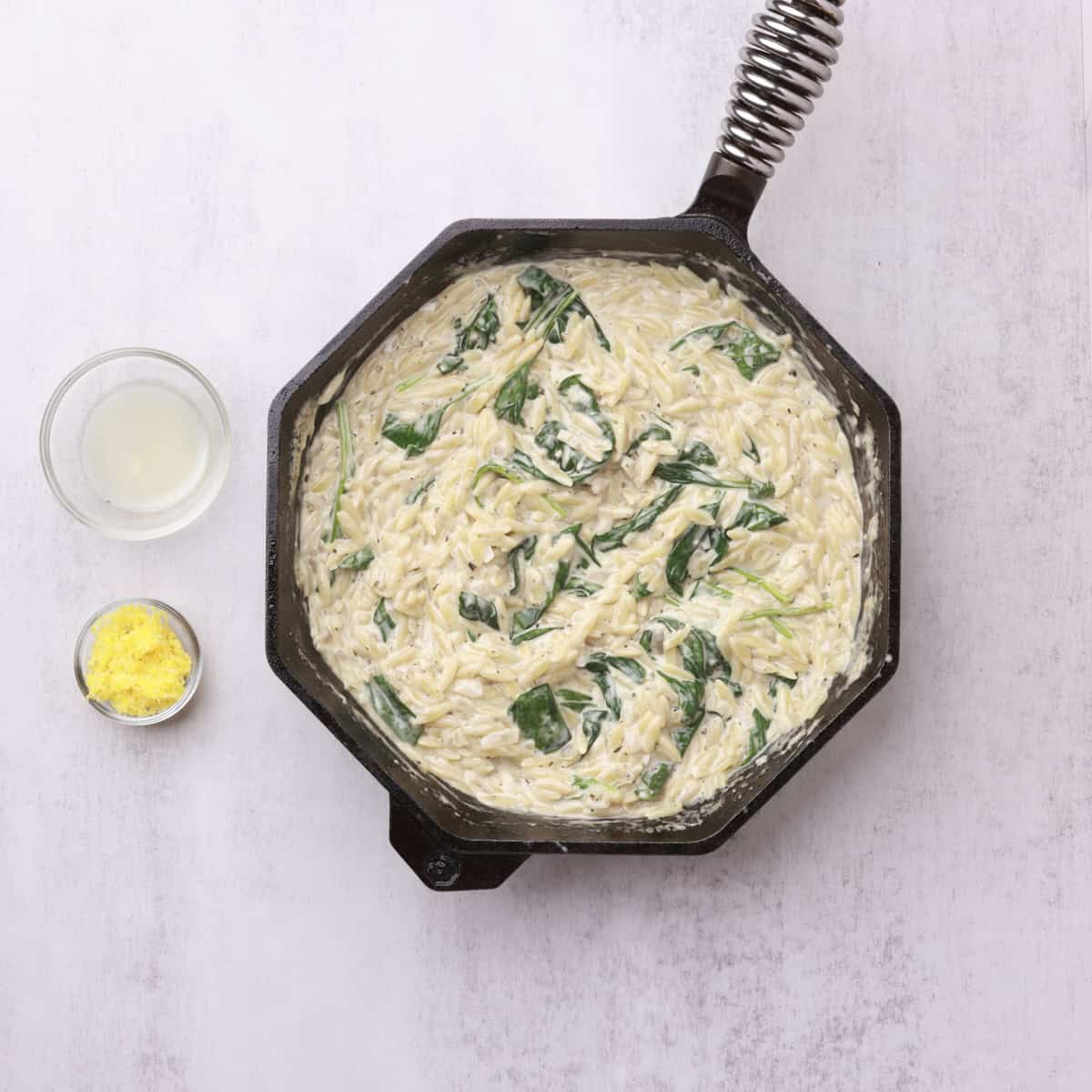 A skillet with the cooked orzo and the cooked spinach in it with the lemon juice and zest in bowls beside.