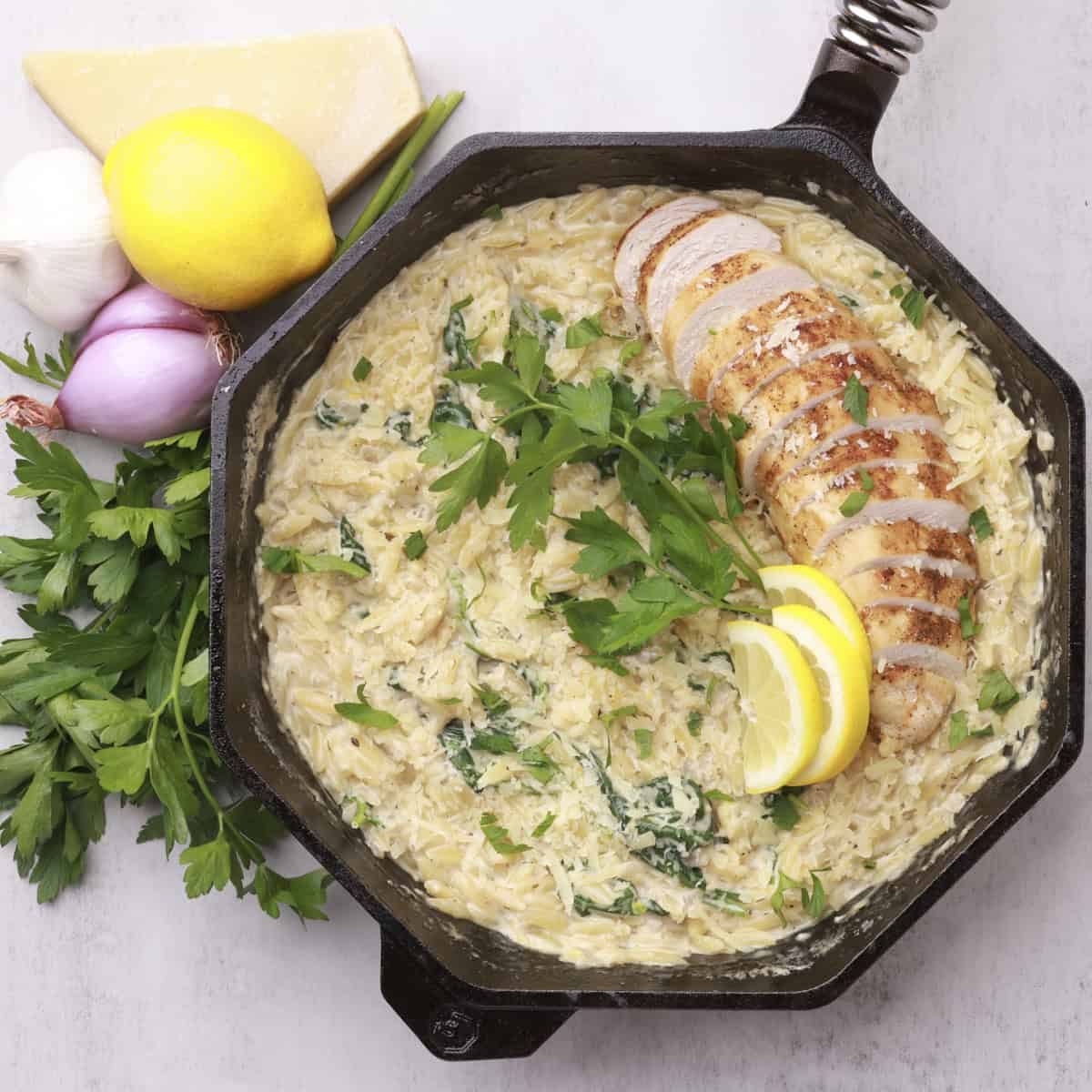 The completed Lemon Chicken Orzo Pasta in a skillet topped with chicken and greens and displayed with more greens and vegetables.