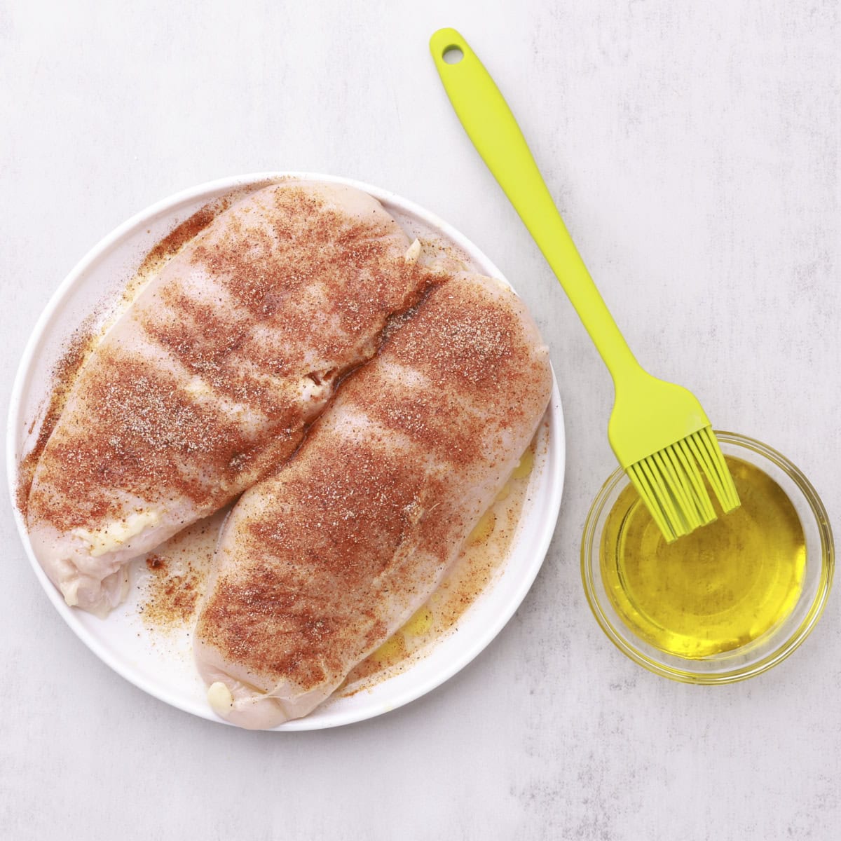 A plate with two seasoned chicken breasts and a small bowl of olive oil with a brush resting on it.