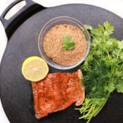 A bowl of Salmon Spice displayed on a cast iron platter with greens, a half lemon and a cooked salmon filet.
