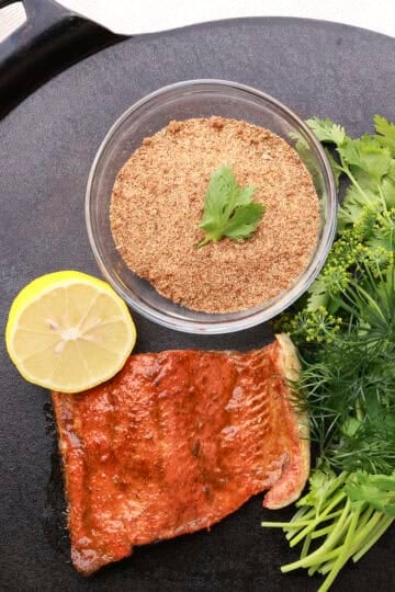 A bowl of Salmon Spice displayed on a cast iron platter with greens, a half lemon and a cooked salmon filet.