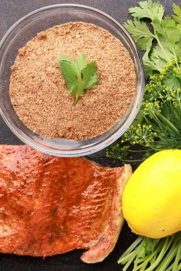 A bowl of salmon spice displayed beside a cooked salmon filet with a lemon and greens.