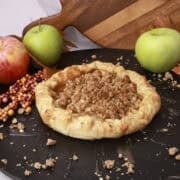 A whole Apple Pie with Crumb Topping displayed on a cast iron sheet with apples in the background.