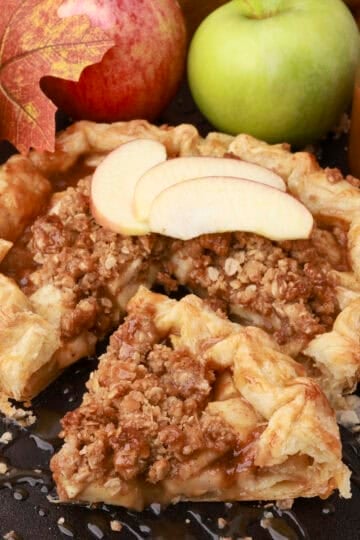 An Apple Pie with Crumb Topping displayed with a slice cut out and displayed in front of it with apples and caramel sauce in the background.