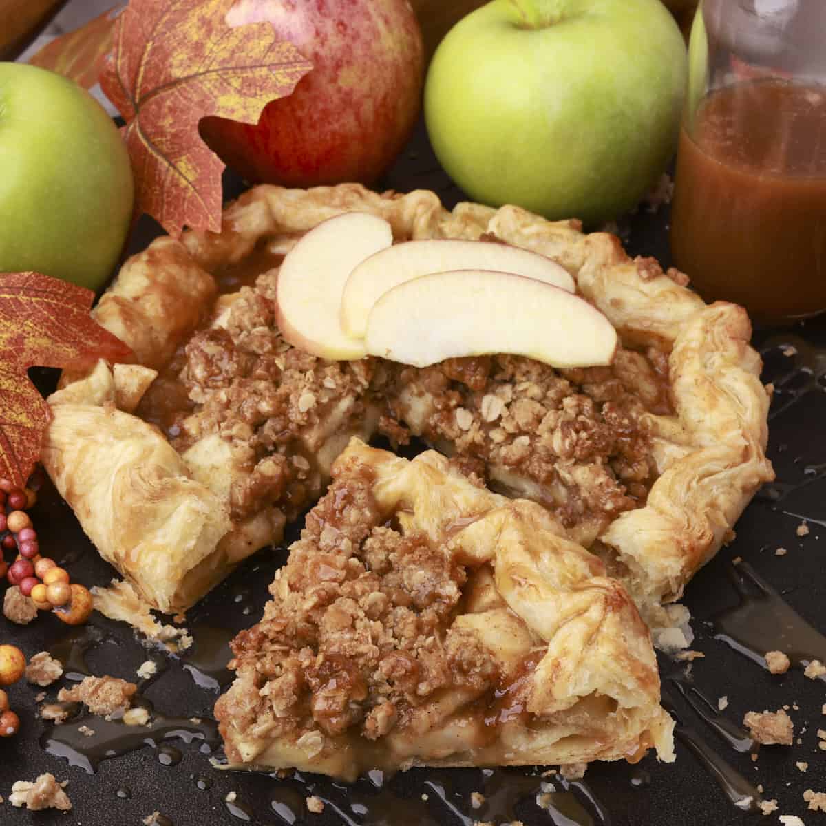 An Apple Pie with Crumb Topping displayed with a slice cut out and displayed in front of it with apples and caramel sauce in the background.
