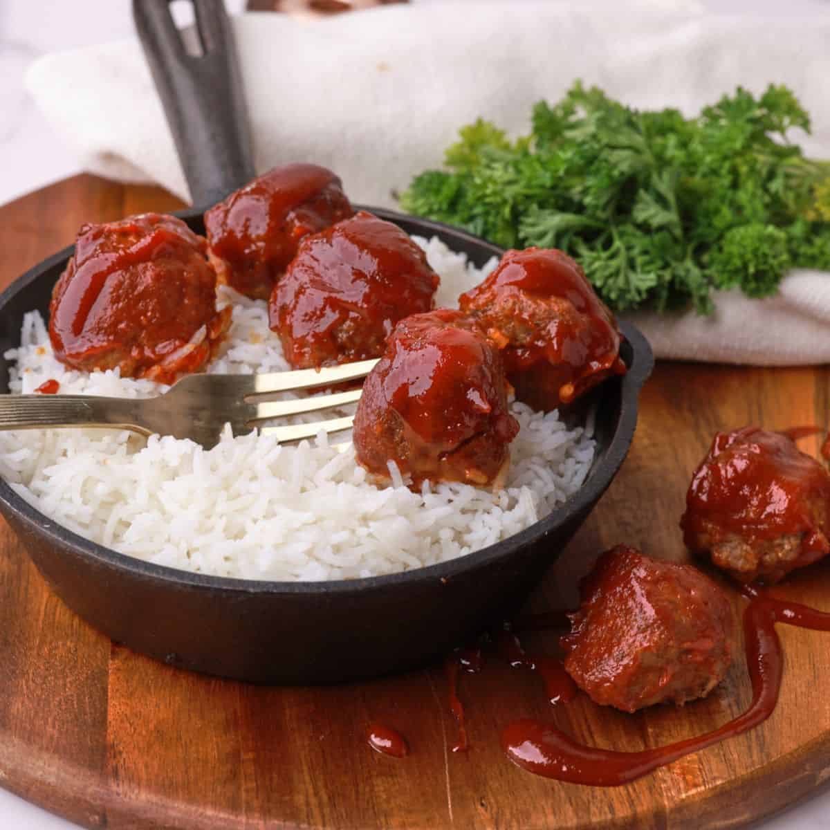 A small skillet filled with rice and topped with meatballs and a fork, displayed on a wooden board with a napkin and greens in the background.
