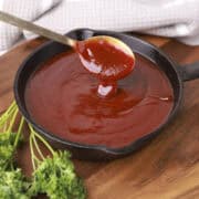 A skillet filled with Homemade BBQ Sauce, displayed on a wooden board with greens and sauce dripping from a spoon above the skillet.