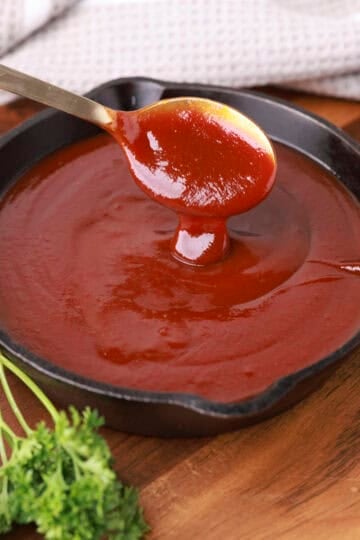 A skillet filled with Homemade BBQ Sauce, displayed on a wooden board with greens and sauce dripping from a spoon above the skillet.